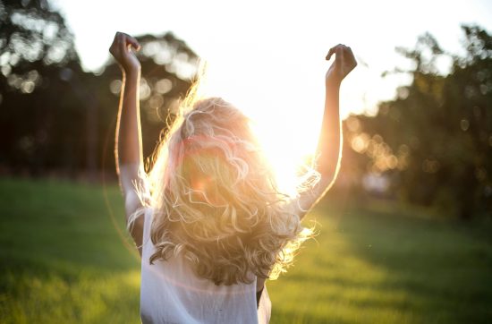 Woman in a green field surrounded by trees and a setting sun with her hands in the air